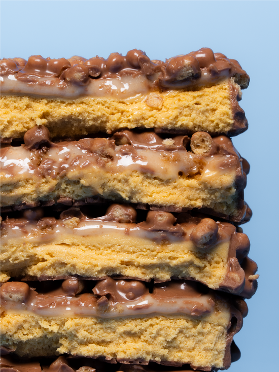 Barebells Cookie Butter protein bar stacked showing layers of golden cake, caramel filling, and chocolate coating with crispy bits against a blue sky background