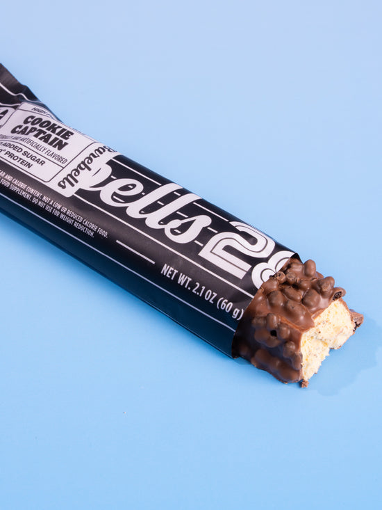Barebells Cookie Captain protein bar with chocolate coating and cookie pieces, partially unwrapped against blue background